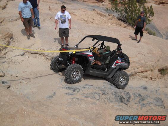 Off-road utility vehicle with a yellow tow strap on rocky terrain, people standing nearby
