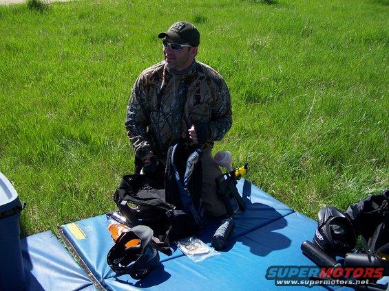 Guy in camo gear kneeling on blue mat with paintball equipment