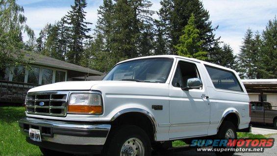 White 1996 Ford Bronco parked in front of a house with trees and a brown pickup truck in the background