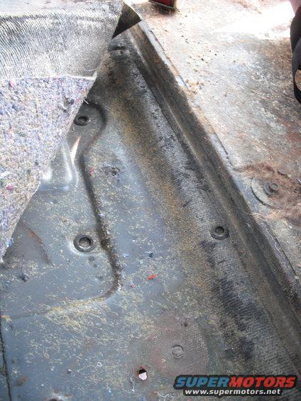 Vehicle floor interior with carpet pulled back, showing metal and mounting points