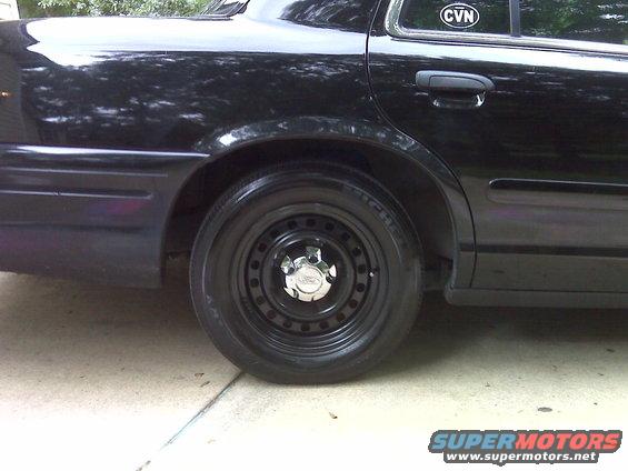 Black sedan's rear wheel with a steel rim and chrome hubcap. Sticker on rear window says CVN
