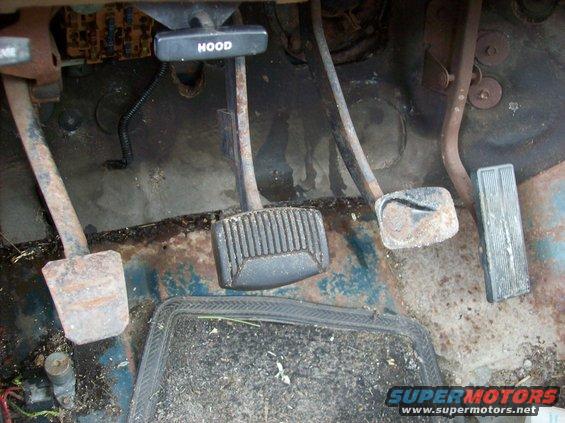 Floor pedals and hood release lever in a vehicle interior. The floor is rusty and dirty