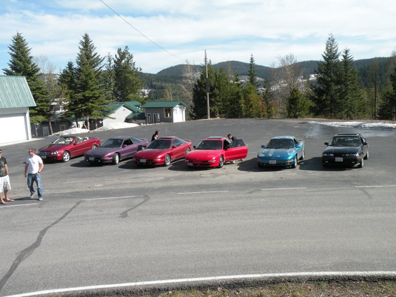 Several sports cars including a red convertible, a purple coupe, two red coupes, a blue coupe, and a black coupe parked in a lot with people standing nearby