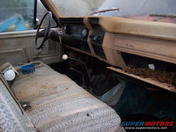 Interior view of a worn pickup truck cab, showing the dashboard, steering wheel, and patterned seat. The glove box is open and filled with debris