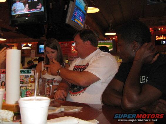 Three people sit at a bar, with TVs on the wall and drinks on the counter