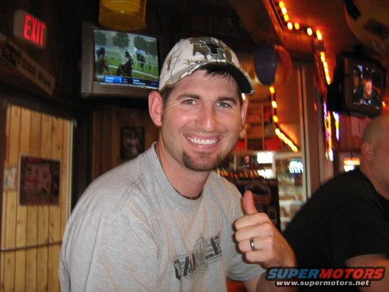 Man in a camouflage hat and grey t-shirt gives a thumbs up in a bar with TVs showing horse racing