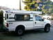 2000 Ford F-250 - A white Ford F-250 pickup truck is equipped with a Grandby camper shell, seen from the side with another white pickup truck in the backgrou…