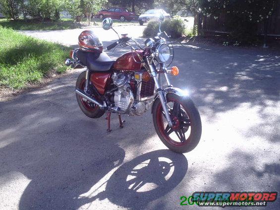 Motorcycle parked on a dirt road, helmet on seat. Two cars in background