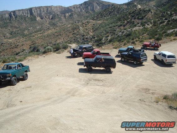 Several off-road vehicles, including a dark SUV and a green pickup truck, are parked on a dusty, mountainous terrain