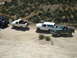 1986 Ford Bronco - Several off-road vehicles are parked on a dusty trail with hills in the background.