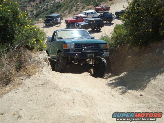 Green 1987 Ford F-150 pickup truck with off-road modifications navigating a steep dirt trail, with other trucks behind it