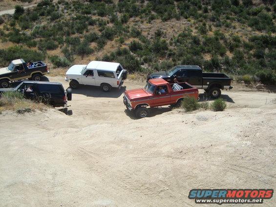 Several off-road vehicles, including a white Bronco and an orange pickup truck, parked on a dirt road