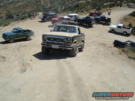 Tan 1980 Ford Bronco with aftermarket wheels and bumper, parked on a dirt road