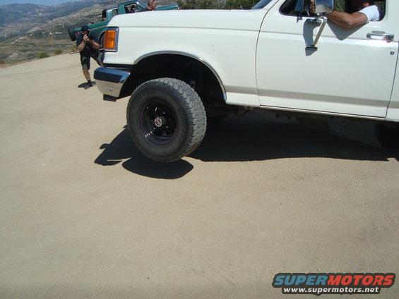 White pickup truck with black wheel, green truck behind it, dusty terrain