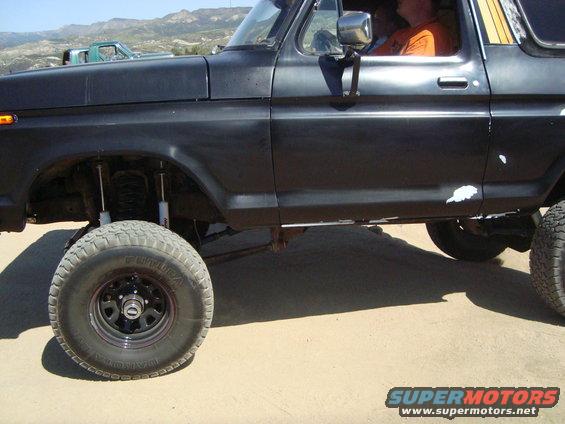 Black 1978 Ford Bronco, lifted with off-road tires, side view. Another green truck is visible in the background
