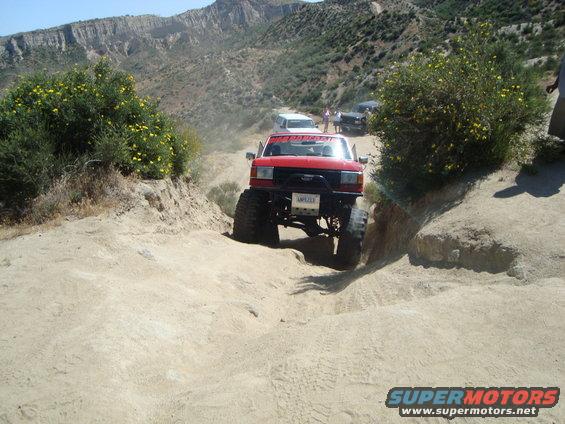 Red SUV with big tires going down a dirt trail. Other vehicles and people are visible in the background
