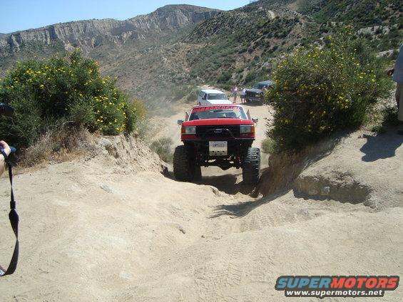Red off-road SUV with large tires driving on a dirt trail, front view. Other vehicles and people are visible in the background