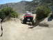 1986 Ford Bronco - A red off-road SUV navigates a dusty trail with other vehicles and people in the distance.