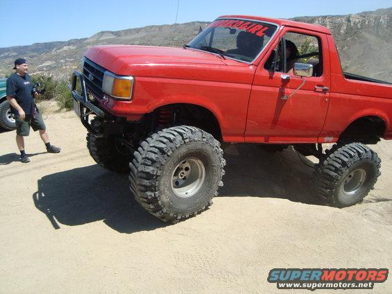 Red lifted Ford Bronco with large off-road tires parked on dirt, person standing nearby