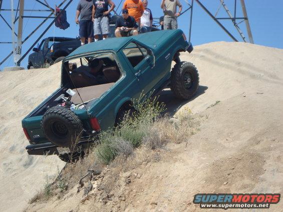Green SUV with spare tire on rear, tilted on sandy incline. Visible interior, off-road tires, and people watching