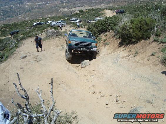 Green SUV navigating a steep, rocky off-road trail with other vehicles parked on the hillside