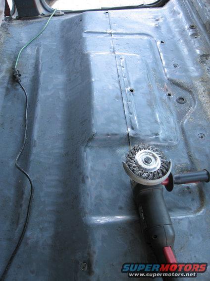 Vehicle floor pan with a wire wheel grinder and a green wire. The floor is gray and shows signs of rust and repair