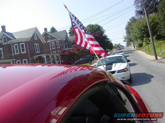 Front of a white 2004 Ford Mustang with a black stripe, an American flag on its roof, in a line of cars