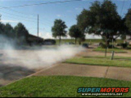 Car doing a burnout, emitting thick white smoke. The vehicle is dark colored and seen from a distance