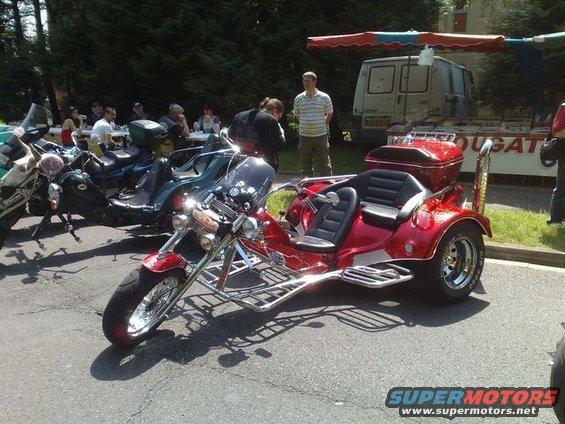 Red trike motorcycle with two seats and chrome accents parked next to other motorcycles