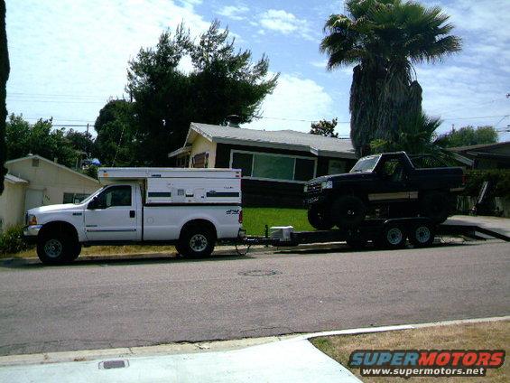 White Ford F-250 with camper shell towing a black Ford Bronco on a trailer