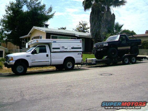 White pickup truck with a camper shell, towing a trailer with a black pickup truck