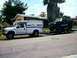 2000 Ford F-250 - A white pickup truck tows a trailer carrying a black pickup truck on a residential street.