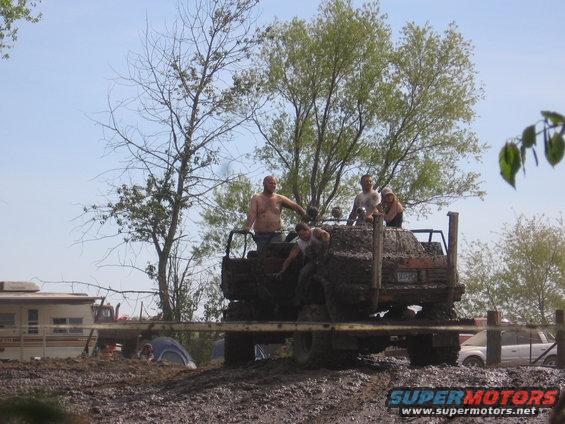 Muddy truck with people riding in the back, stuck in the mud. Another vehicle is visible in the background