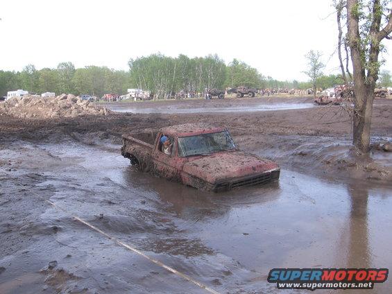 Red pickup truck stuck deep in mud, with other vehicles and spectators in the background