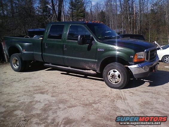 Green Ford F-350 Super Duty pickup truck with dually rear wheels parked on dirt. White car visible in background