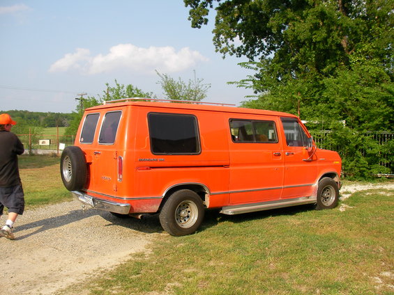 Orange Ford E150 van parked on grass with a spare tire on the back. A person in an orange hat stands nearby