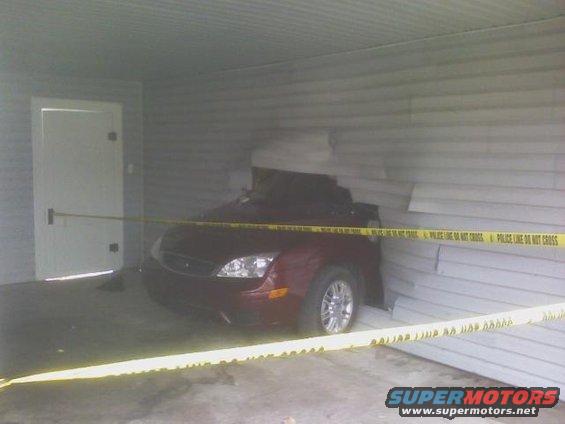 Red sedan with front end damaged, stuck in a building wall. Police line tape is across the scene