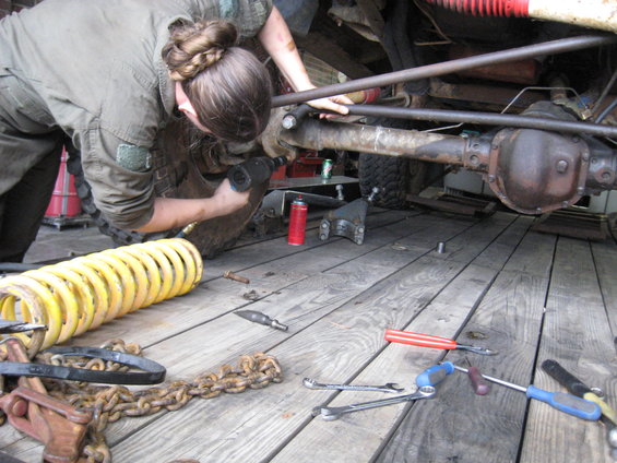 Person with braided hair using an impact wrench on a vehicle's rear axle and suspension components. Yellow coil spring and chains are on the wooden floor