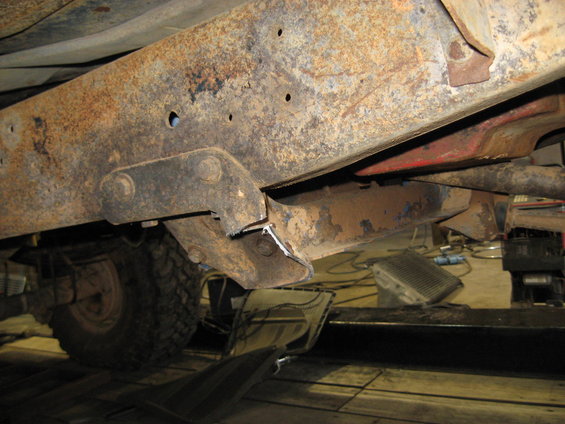 Heavily rusted vehicle frame with visible suspension parts and a tire. The vehicle is on a lift