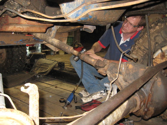 Man wearing glasses and a blue shirt works on the driveshaft and differential of a rusty vehicle