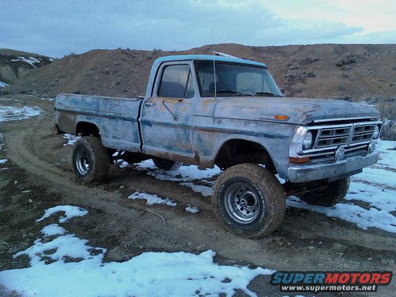 Blue 1970 Ford F-100 pickup truck with lifted suspension and off-road tires, parked on a dirt road with patches of snow