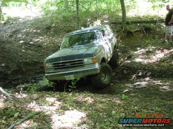 Ford Bronco with lifted suspension navigating muddy terrain in a wooded area. License plate reads 2S2W024