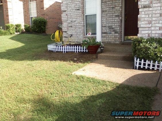 Front yard with a brick house, walkway, and some landscaping. A yellow hose is coiled near a small white picket fence