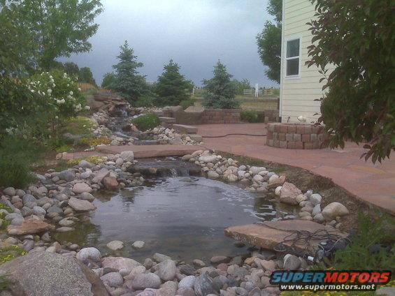 Backyard scene with a pond, waterfall, stone patio, and a house