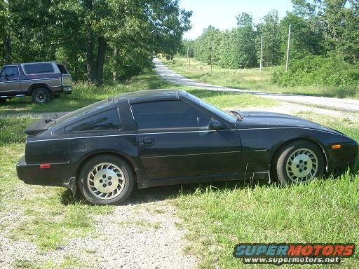 Black 1989 Nissan 300ZX parked on grass next to a dirt road; older Ford Bronco visible in background