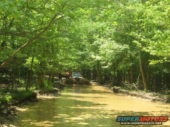 Two off-road vehicles splashing through a muddy stream in a dense forest