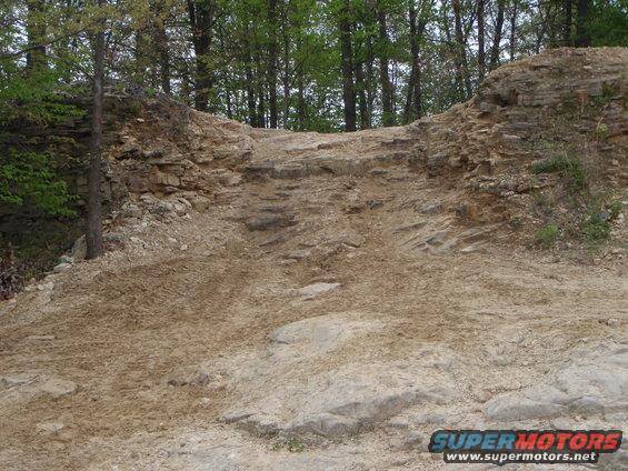 Off-road trail with rocky terrain and trees in the background. Supermotors.net logo in lower right