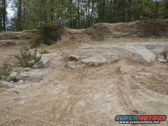 Sandy off-road trail with tire tracks and rocks, leading up a dirt hill with trees in the background