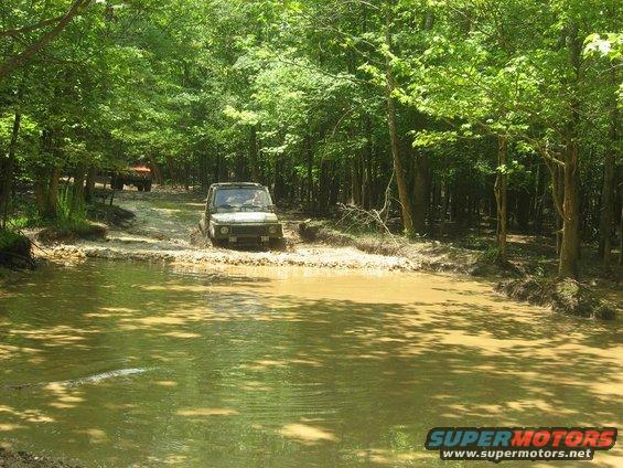 Dark green 1987 Suzuki Samurai driving through a muddy water crossing, with another off-road vehicle behind it on a dirt path
