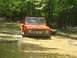1987 Suzuki Samurai - Orange Ford Bronco navigating a mud pit on a wooded trail.